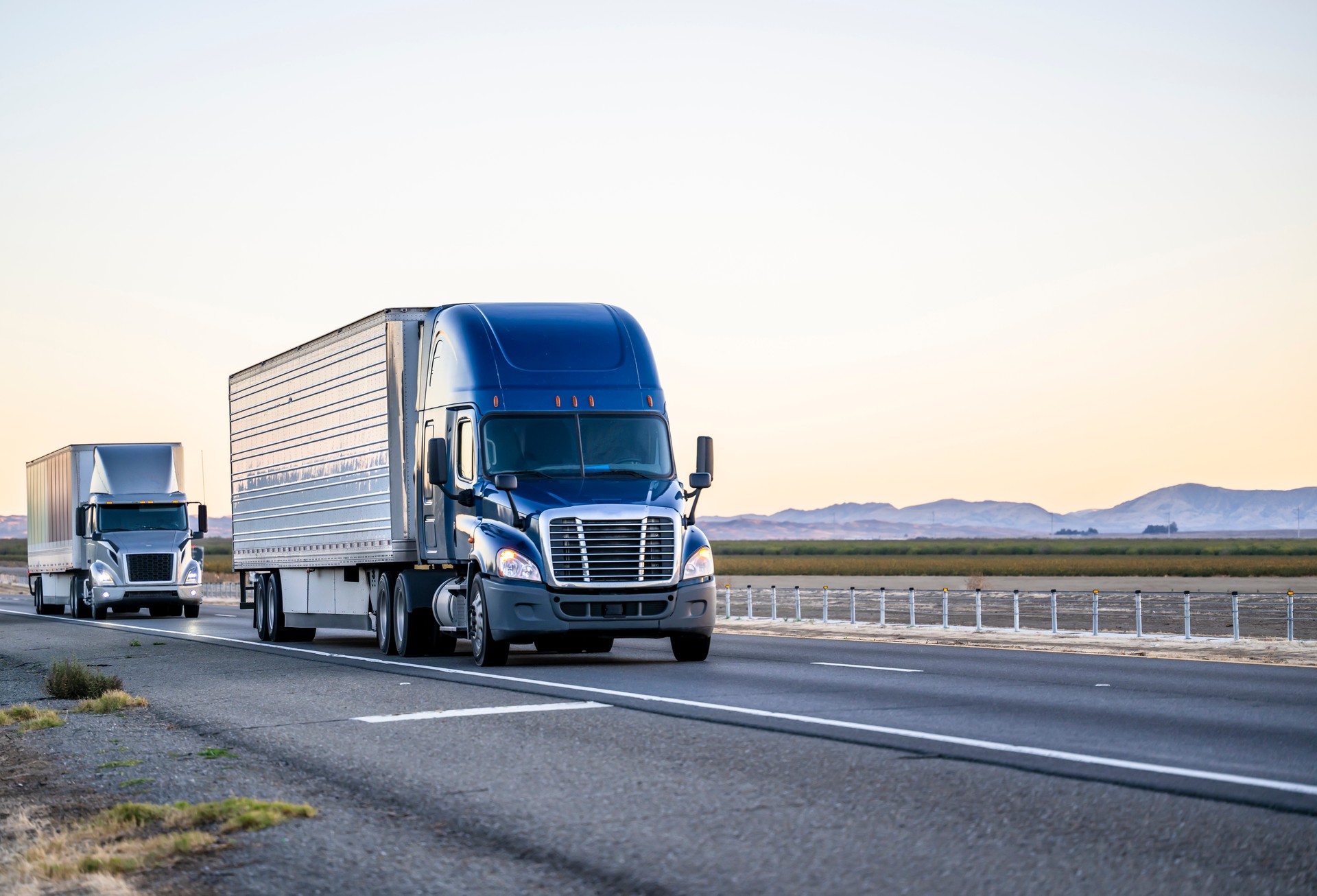 Two industrial bonnet big rigs semi trucks transporting cargo in different semi trailers driving on the straight highway road in California